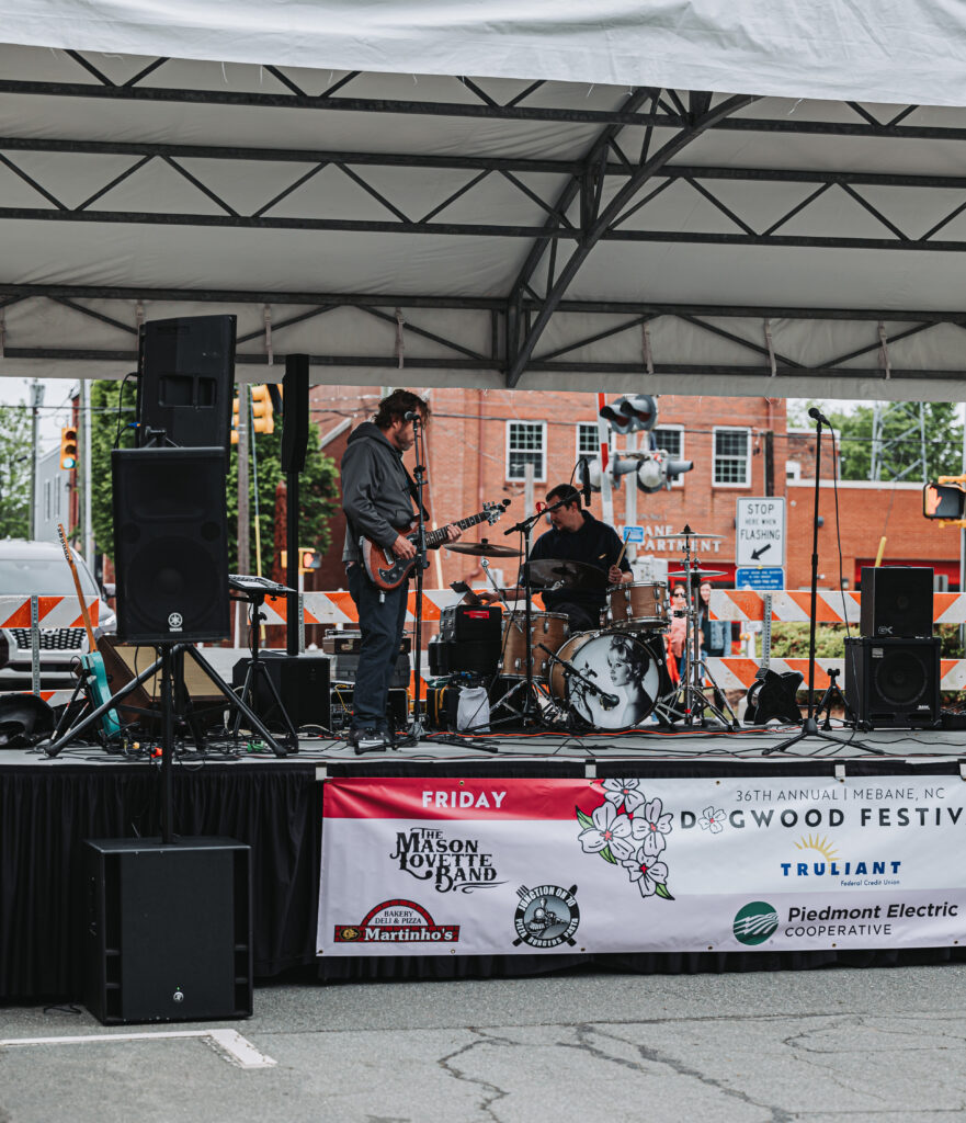 image of a music stage with guitarist ad drummer warming up for the nightly performance. there's a banner at the front of the stage with dogwood festival written on it and sponsors of the event