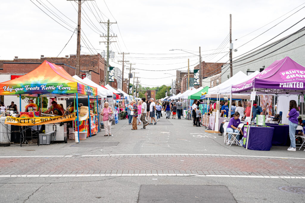 image of downtown mebane with vendors lining the streets selling goods, food, or having arts and crafts like face painting. many people roaming the downtown streets checking out the vendors.