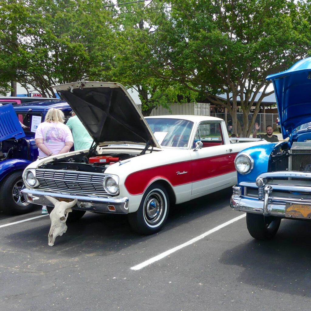 image of white truck with red stripe in parking lot at car show with its hood open and an animal skull at the front of it with a slight view of a blue vehicle with its hood open