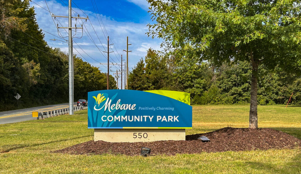 Mebane Community Park sign on blue and green surrounded by green trees and bright blue skies. 