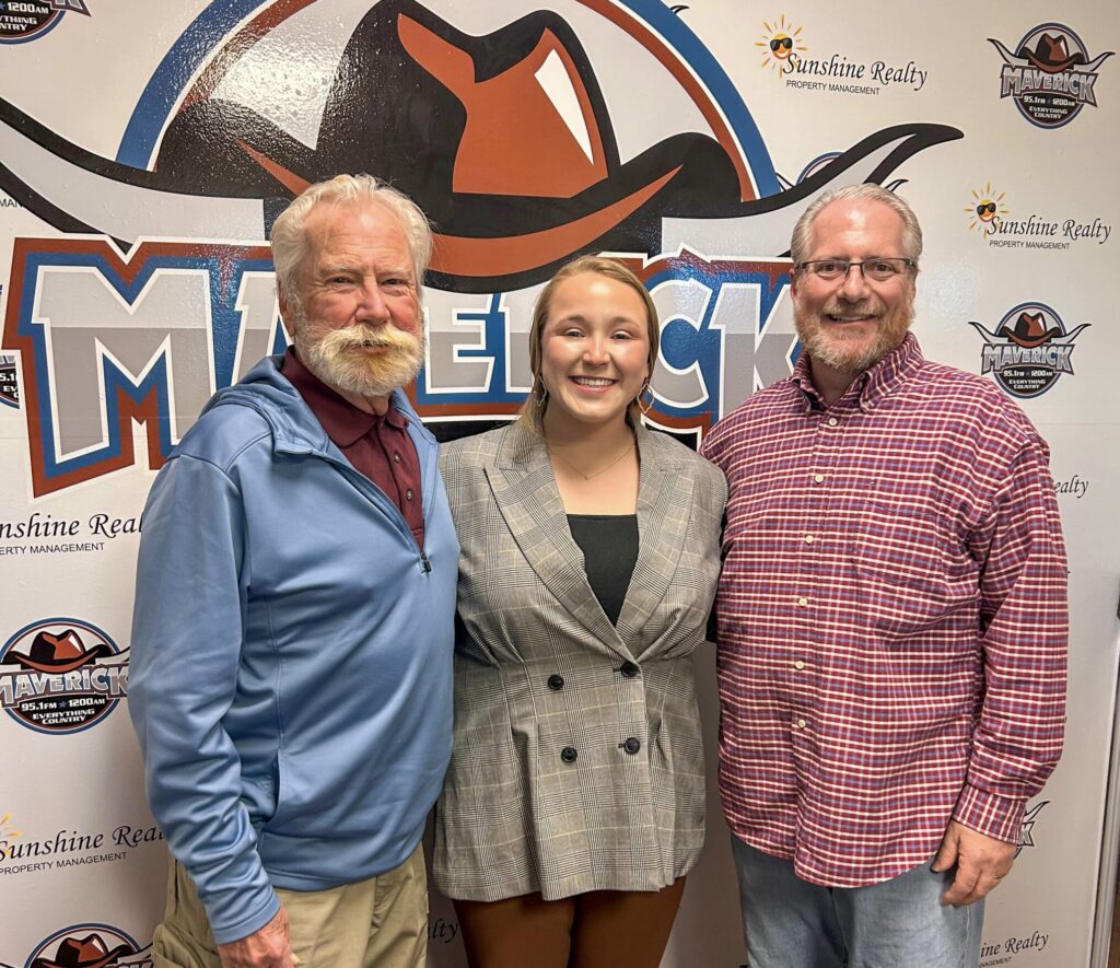 Group of two men and one woman standing side by side in front of a wall decorated with Maverick cowboy hat logo and Sunshine Realty text.