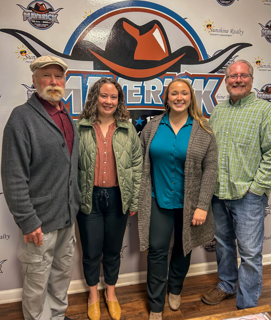Four adults standing indoors in front of a Maverick-themed backdrop with logos, dressed in casual and business casual attire.