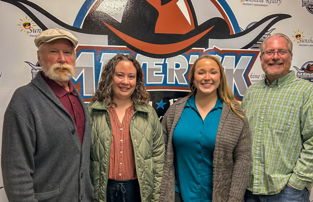 Four adults standing indoors in front of a Maverick-themed backdrop with logos, dressed in casual and business casual attire.