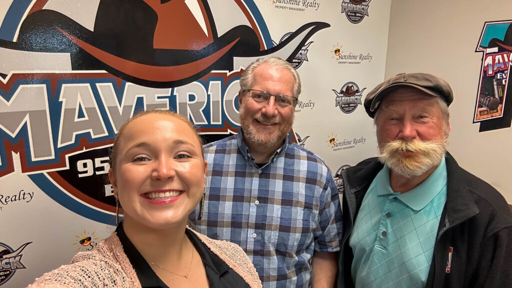Three people standing in front of a Maverick 95.1 FM radio station logo wall with promotional posters around them.