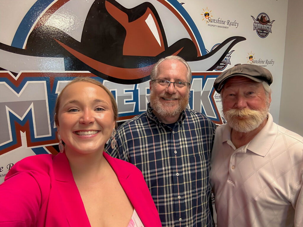 Group selfie of two men and one woman standing in front of a wall decorated with a large cowboy hat logo and the word 'Maverick'.