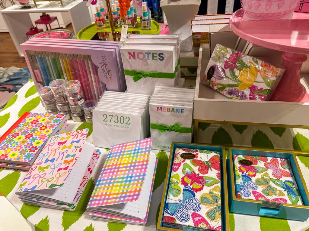 Image of Pink Lola's stationary table filled with colorful notepads and some marked with Mebane and pins and patterned napkins. 
