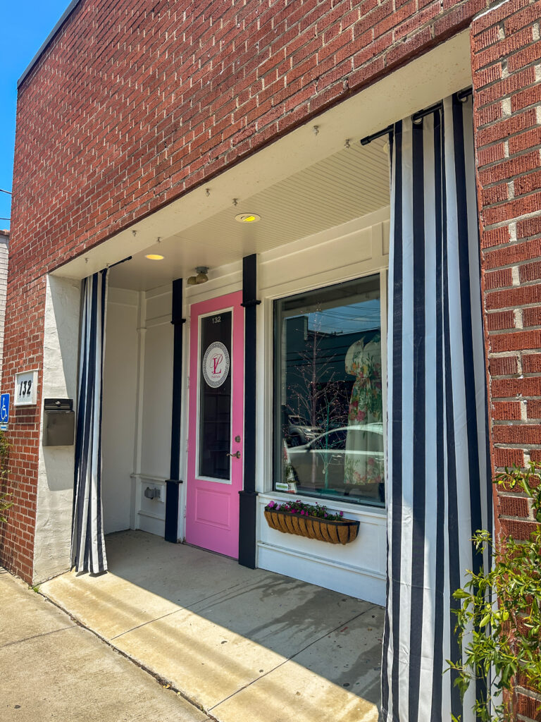 Outdoor view of the store Pink Lola. The storefront has a pink door and black and white striped curtains. 