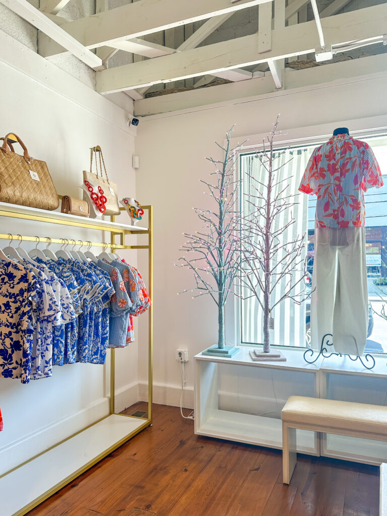 Inside view of Pink Lola showing the window display of mannequin dressed in Pink Lola's clothes. The Image also shows a clothing rack with blue shirts lined up and purses sitting atop the rack.