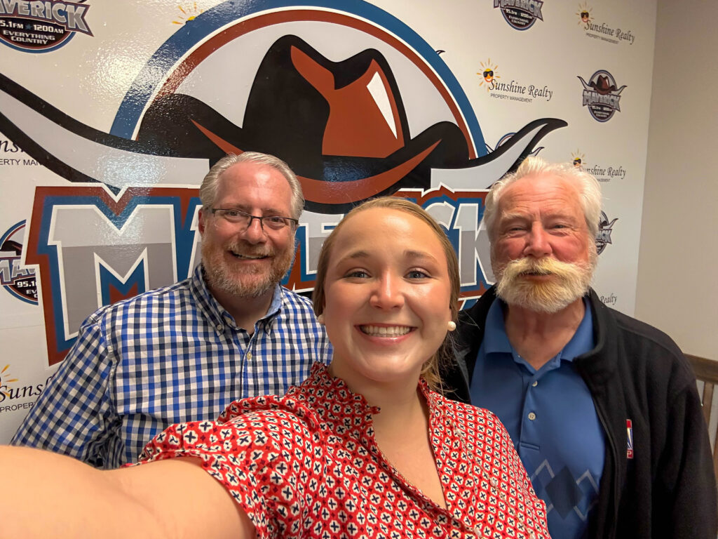 Three people posing for a selfie in front of a wall with a large Maverick logo featuring a cowboy hat and horns.