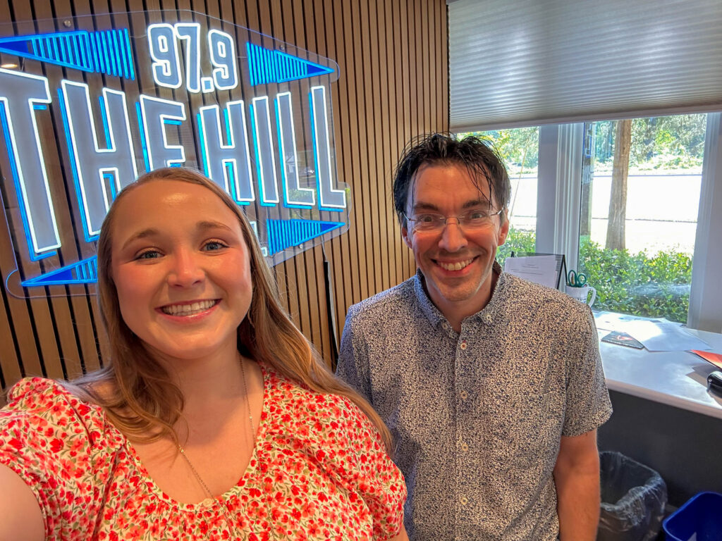 Radio station interior with two people smiling near a window and a prominent 97.9 THE HILL sign on a wooden wall behind them. 
