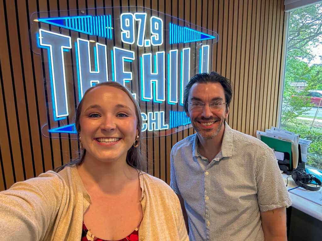 A man and woman stand inside a radio station studio with a glowing 97.9 THE HILL WCHL sign on a wooden slat wall behind them.